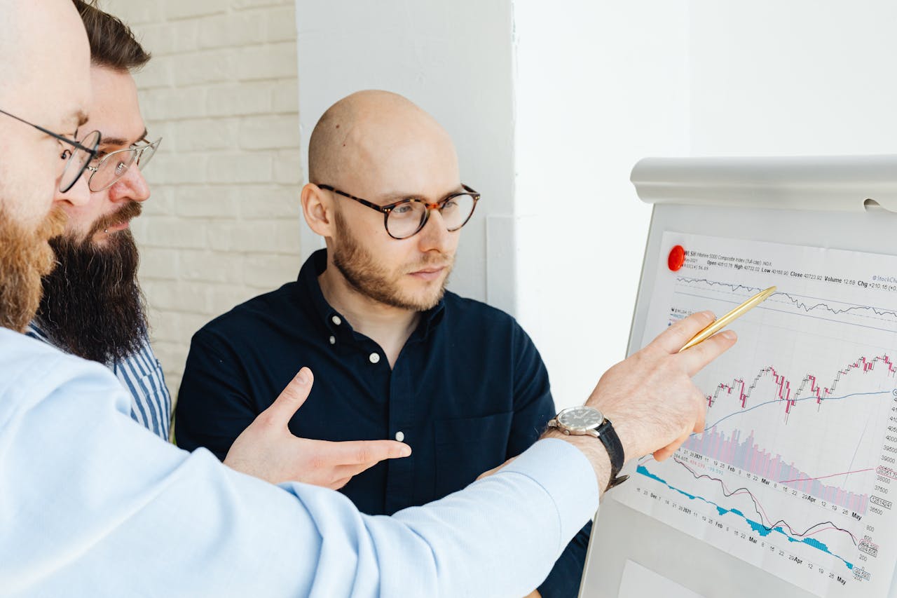 Contact Three men discussing financial charts on a whiteboard during a business meeting.